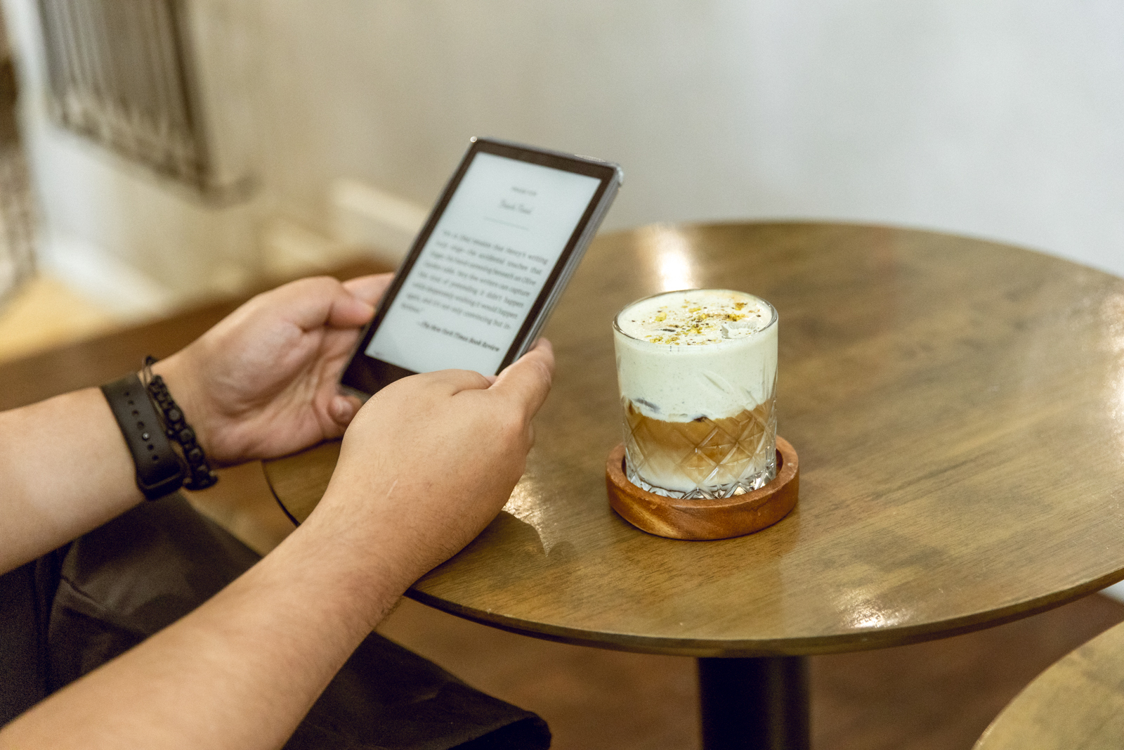 Photograph showing a person holding an e-reader while sitting at a round wooden table with a glass of layered iced coffee topped with cream and sprinkled garnish. The scene captures a relaxed moment in WHAT ABOUT COFFEE, in Katipunan, Quezon City, combining digital reading and a specialty beverage in a cozy indoor setting.