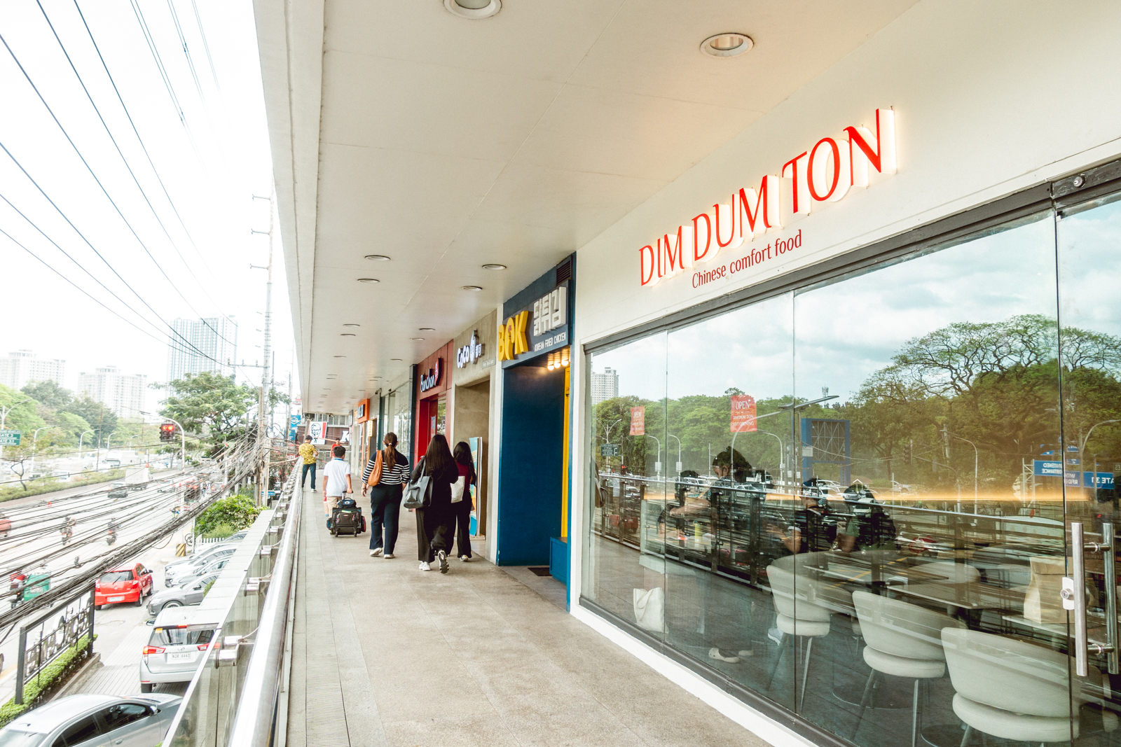 Photograph of an outdoor shopping walkway featuring several storefronts at Regis Center, Katipunan, Quezon City, including a prominently visible restaurant named "DIM DUM TON" with a tagline "Chinese comfort food." The scene includes pedestrians walking along the corridor, a glass window reflecting street traffic and trees, and a multi-lane road with vehicles on the left side.