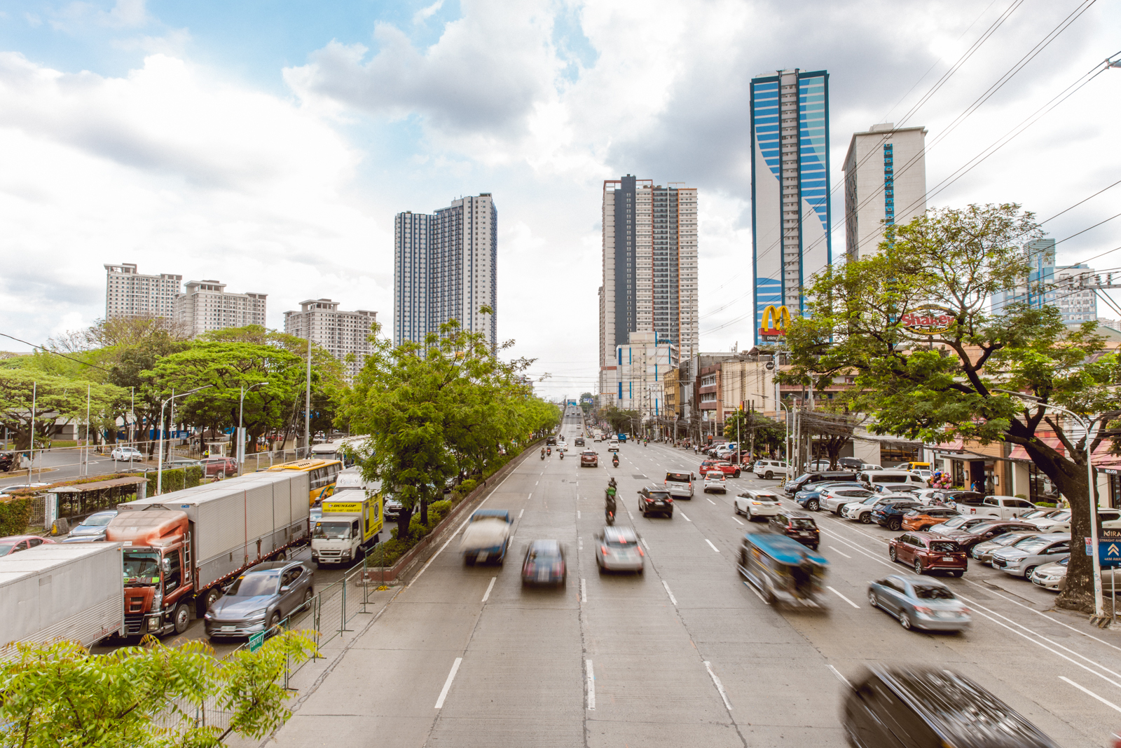 Photograph showing a busy Katipunan avenue with multiple lanes of moving vehicles, including cars, trucks, and motorcycles, flanked by trees and parked cars.