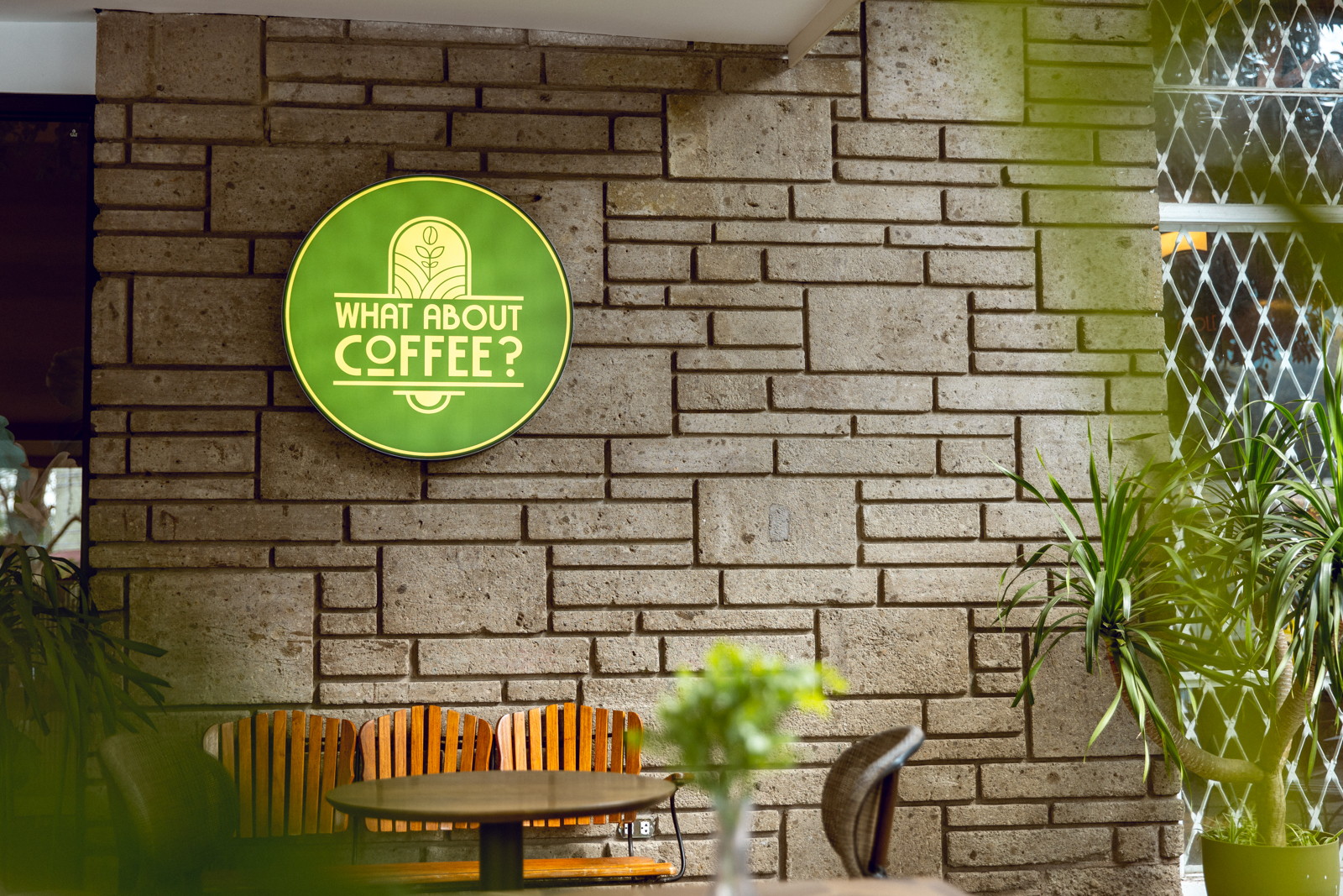 Photograph of a coffee shop exterior featuring "WHAT ABOUT COFFEE?" signage in Katipunan, Quezon City, mounted on a textured brick wall. The setting includes wooden chairs, a round table, and several green plants, creating a cozy and inviting atmosphere.